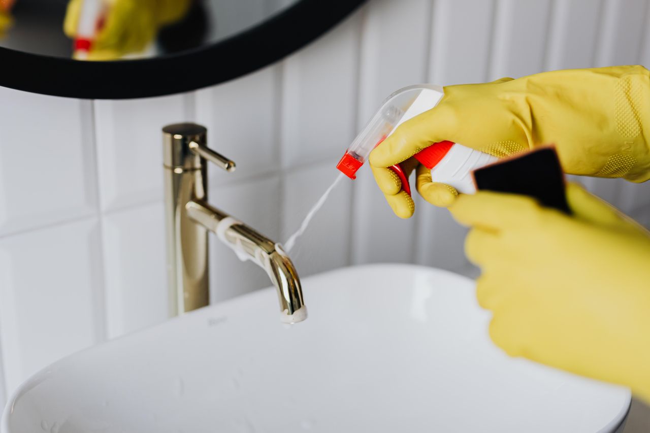 Seen from above, an anonymous housekeeper wearing yellow rubber gloves sprays cleaner onto a chrome bathroom water faucet.